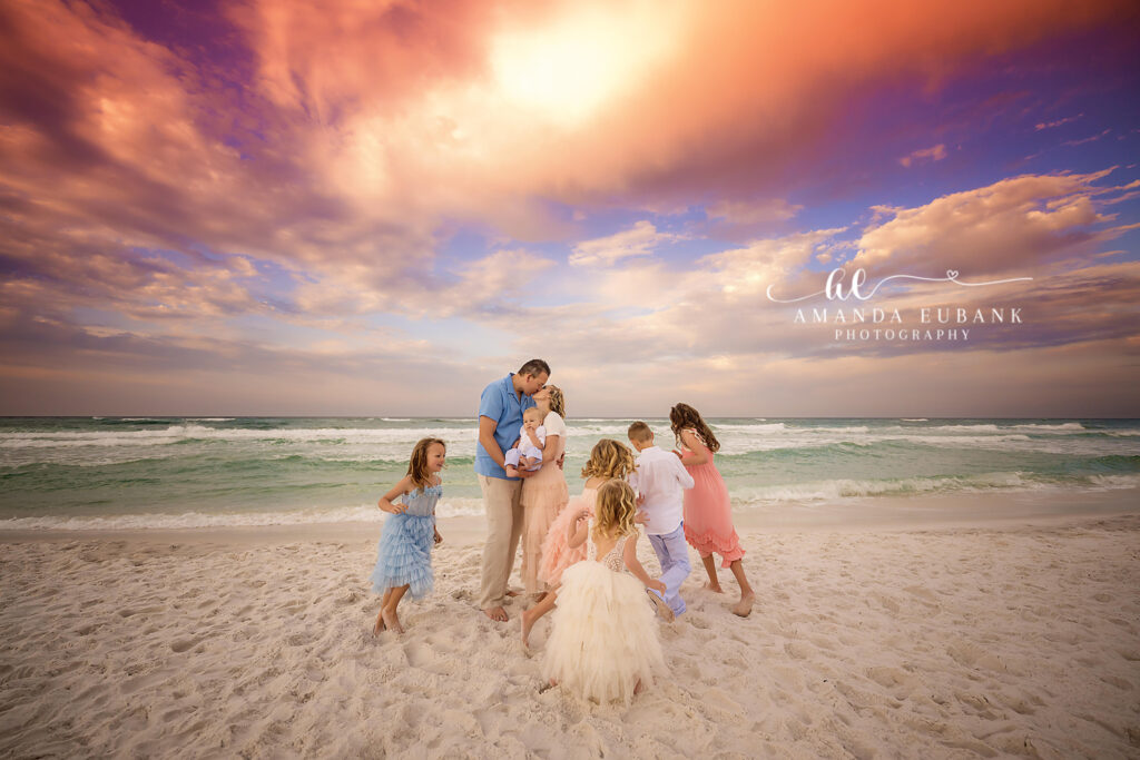Large coordinated family — parents and six children in pastel pink, blue, peach and coral — mom with the four daughters on Santa Rosa Beach at warm-orange sunlit horizon.