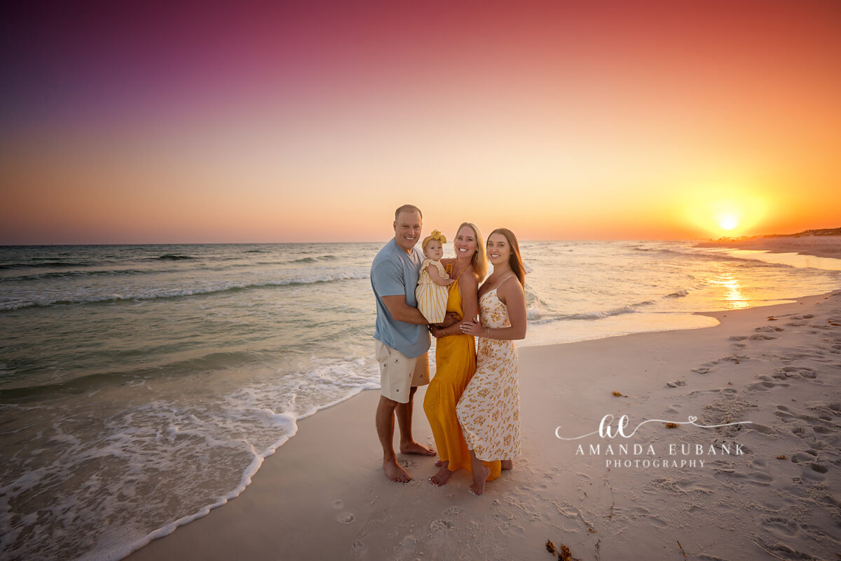 Seaside Florida Family Sunset 198 — wide family environmental shot at rose-tinted twilight Coordinated family of three — mom in a mustard halter maxi, dad in soft blue, and their baby girl in a cream-and-yellow striped sundress — wide family environmental shot along Seaside, FL at rose-tinted twilight.
