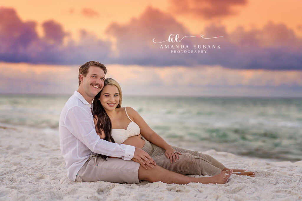 A romantic maternity couple pose at the shoreline against a glowing Seaside sunset sky during crimson and gold dusk.