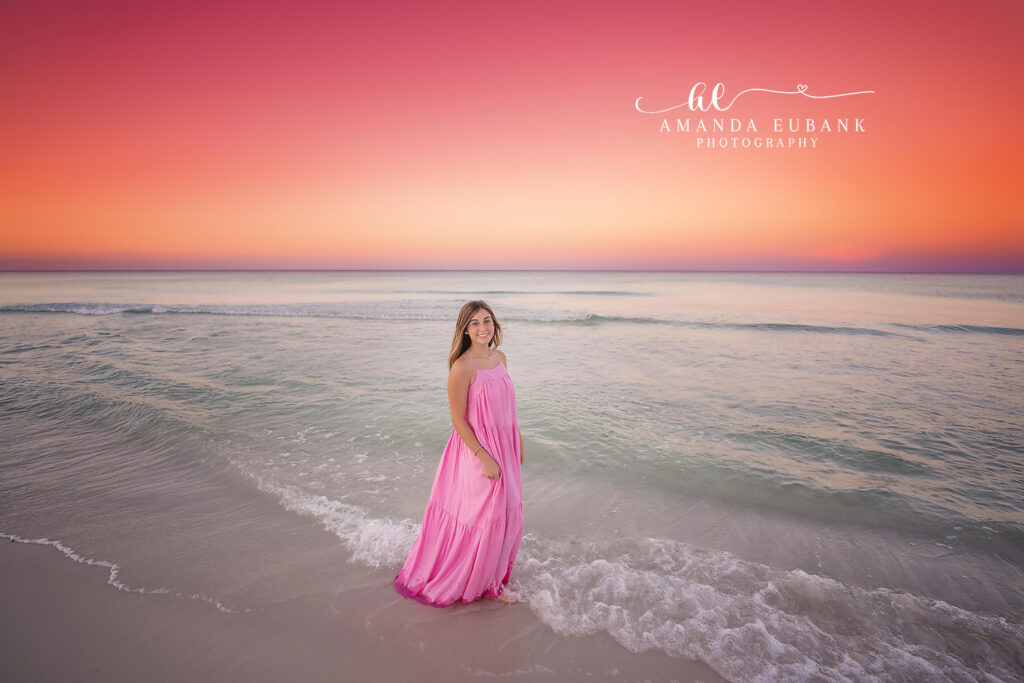 A serene Seaside senior pose in pink as the sky blushes peach and gold during sun-drenched afternoon.