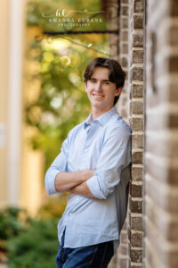 High school senior boy in a light blue button-up leaning casually against a sunlit brick wall with arms crossed
