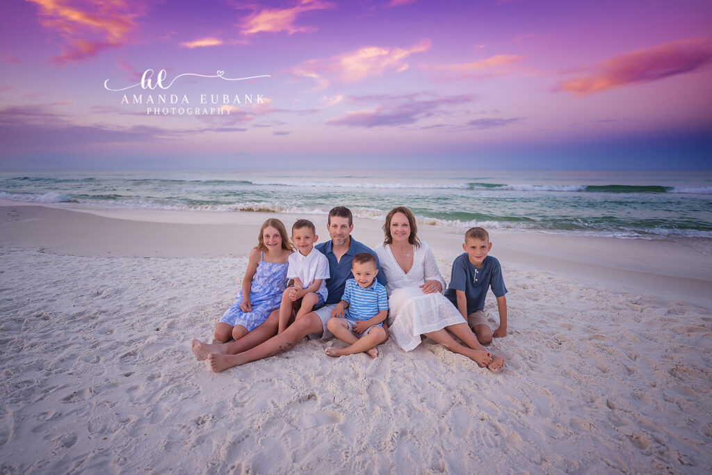 Cannon family of six seated together in the soft beach sand in a white eyelet maxi, blue floral dress, and boys in coordinated blue and white at sunrise on 30A.