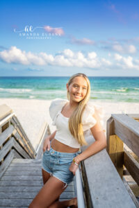 a senior leaning on a weathered beach boardwalk railing in a white flutter-sleeve crop top with denim cutoffs at a sunlit beach access boardwalk along 30A, a clear blue mid-day sky with scattered cumulus clouds.