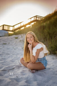 the senior seated in the sand with a sunset glow behind her in a white flutter-sleeve crop top and denim cutoffs at the powdery beach at golden hour, a glowing tangerine sunset sky.
