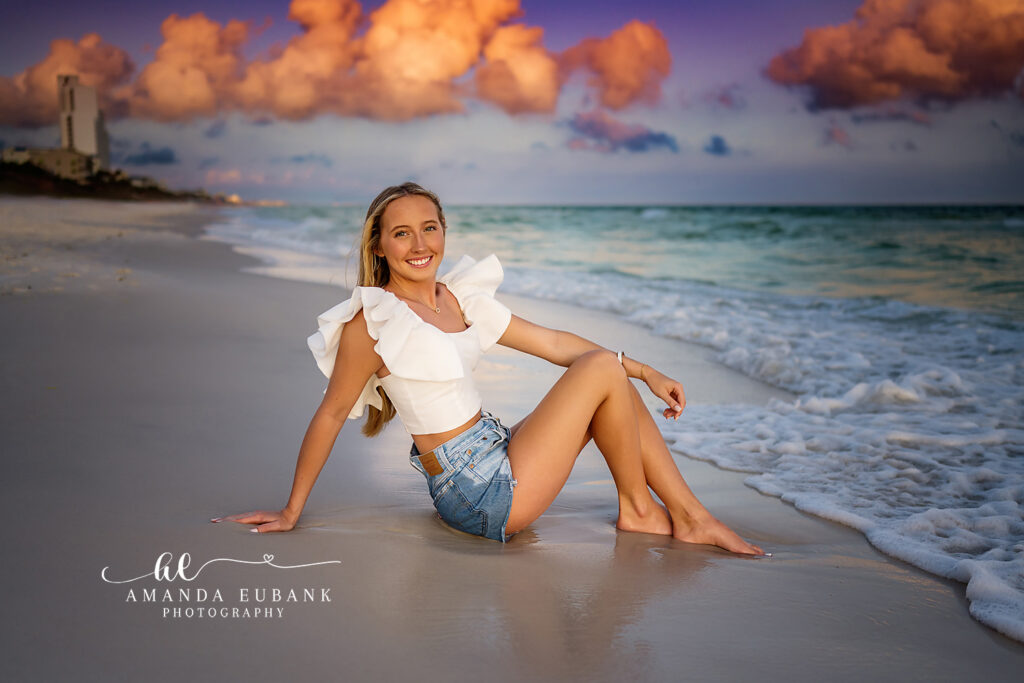 the senior posing solo at the water line in a white crop top and denim cutoffs at the gulf shoreline at 30A, a rich golden sunset sky.