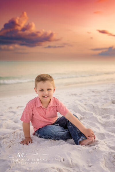 Miramar Beach Family Photographer | Younger Brother in Coral Polo Young boy in a coral polo and denim jeans seated in the powdery shoreline sand at Miramar Beach, Florida beneath a pastel pink-and-orange sunset