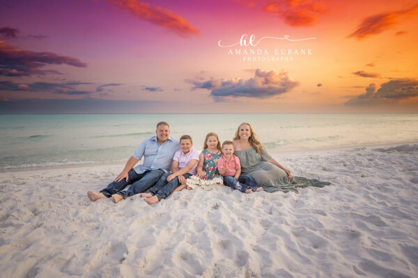 Miramar Beach Family Photographer | Family of Five Sunset Pose Young family of five at the water line in coordinated sage green, coral, white, and denim along the emerald Miramar Beach shoreline beneath a vibrant pink-and-orange sunset