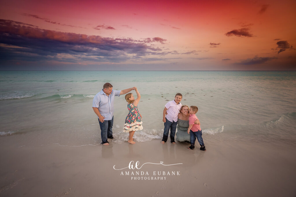 Dad in a light blue shirt twirls his daughter in a teal floral dress as her brothers cheer in the Gulf surf at Miramar Beach beneath a vivid pink-and-orange sunset