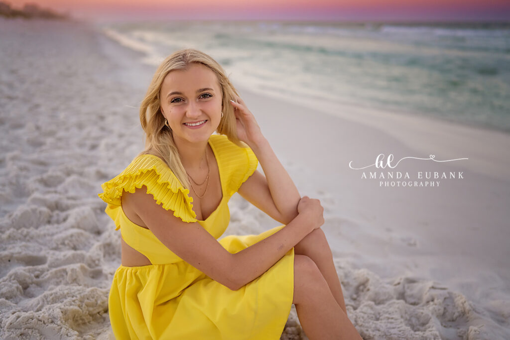 Blonde high school senior in a yellow ruffle dress seated on white sand with the emerald Gulf and pastel sunrise behind her on 30A