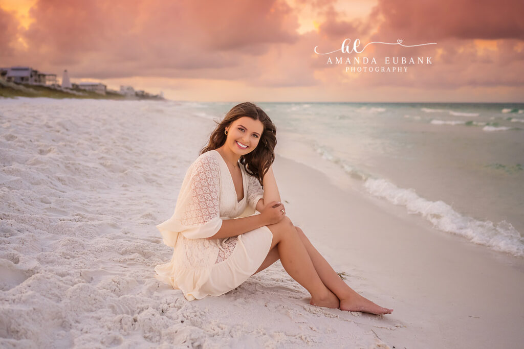 Watercolor sunrise senior Miss R seated on the sugar-white sand at sunrise as peach and pink clouds glow above the Watercolor beach houses behind her, in cream and ivory bohemian lace dress with bell sleeves | Watercolor FL Sunrise Senior Portrait Photographer