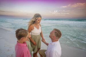 Tender family moment as dad kneels and mom leans toward the kids beside the jade Gulf waves at Destin under a violet-and-cream sunset sky.