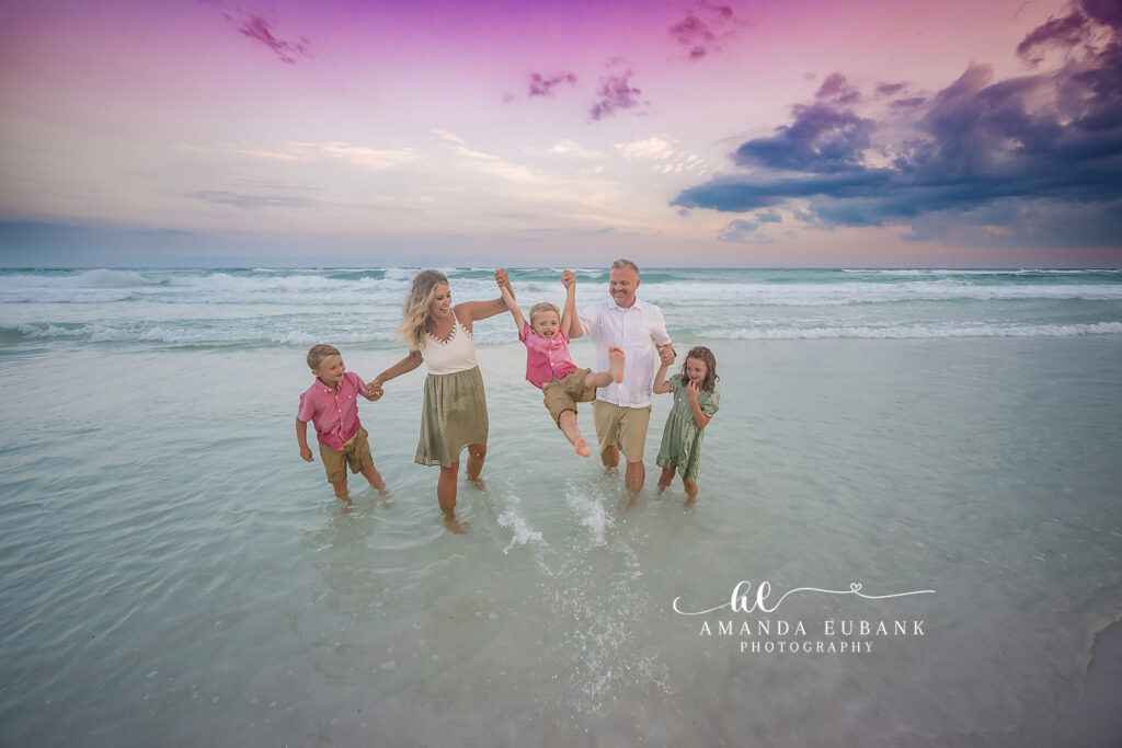 Family of five standing in shallow Gulf surf at Destin while mom and dad swing the youngest boy by his hands, his older brother and sister laughing beside them under a violet sunset.