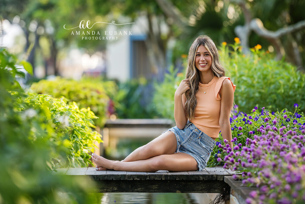 Smiling senior in peach tank and denim shorts seated on the wood ledge above a Watercolor koi pond, lush blooms and lantana cascading around her.