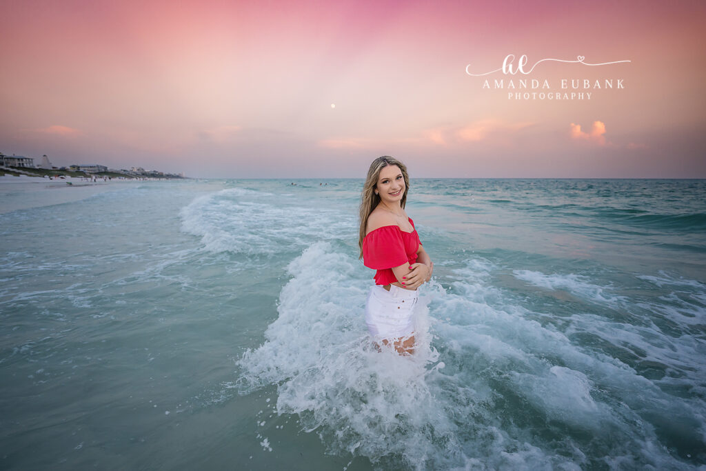 Watercolor senior Peyton standing in the surf with the coral peplum catching breeze against the deepening twilight sky, in coral off-shoulder peplum crop top and white denim shorts | Watercolor FL Senior Portrait Photographer