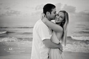 Black and white portrait of a newly engaged couple embracing as the groom kisses the bride on the cheek at Santa Rosa Beach