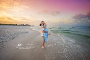 Groom lifting his bride to be in the air as they kiss in the surf at sunset on 30A with a vivid pastel sky overhead