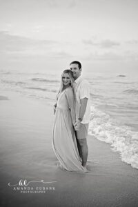 Black and white full-length portrait of a newly engaged couple standing on the wet sand of Santa Rosa Beach with the bride in a flowing gown