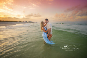 Newly engaged couple standing waist-deep in the emerald Gulf with foreheads touching beneath a vibrant pastel sunset on 30A