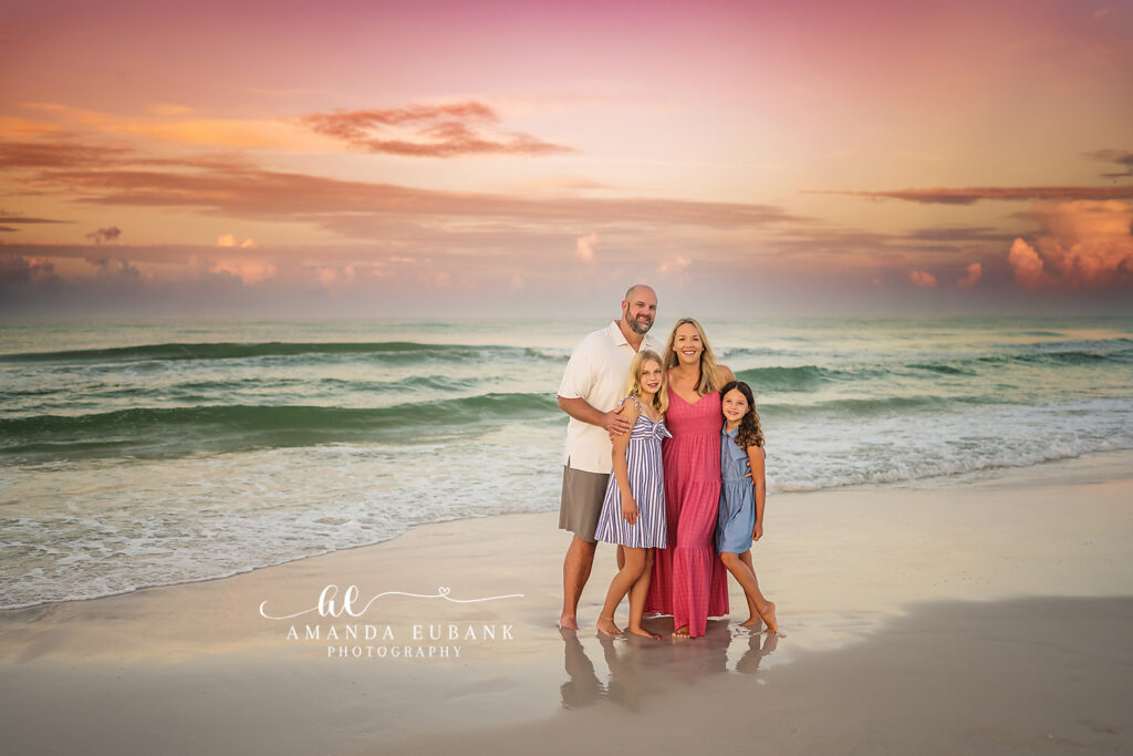 Family of four standing on the mirror-like wet sand at Seaside, sunrise reflections glowing beneath their bare feet under a lilac sky.