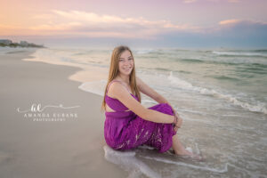 Senior girl in fuchsia maxi seated with knees pulled up at Watercolor shoreline edge with surf rolling at her feet at soft pink dawn.