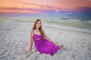 Senior girl in fuchsia maxi seated relaxed in Watercolor sand at painted lavender and peach pre-dawn sky.