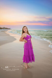 Vertical full length senior portrait of girl in fuchsia maxi standing on Watercolor shoreline beneath painted lavender and peach pre-dawn sky.
