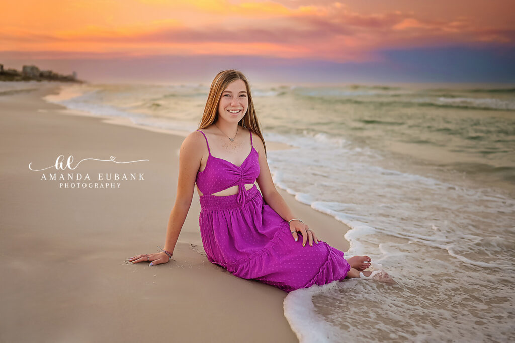 Horizontal senior portrait of girl in fuchsia maxi seated relaxed at Watercolor shoreline edge with surf rolling at painted golden peach pre-dawn.