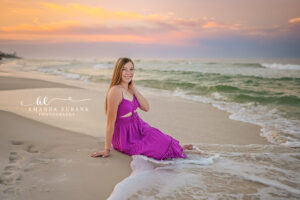 Horizontal senior portrait of girl in fuchsia maxi seated at Watercolor shoreline edge with hand to cheek beneath painted peach and lavender pre-dawn.