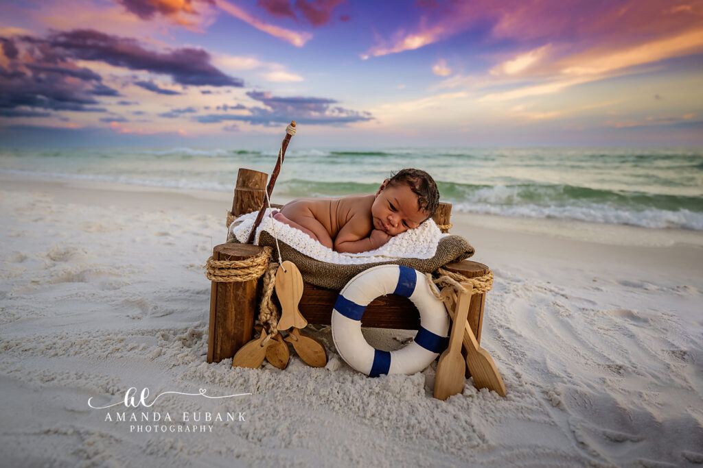 Newborn baby boy sleeps curled inside a small wooden boat with a life ring on the Destin sand as a purple, orange, and blue sunset sky glows behind him.