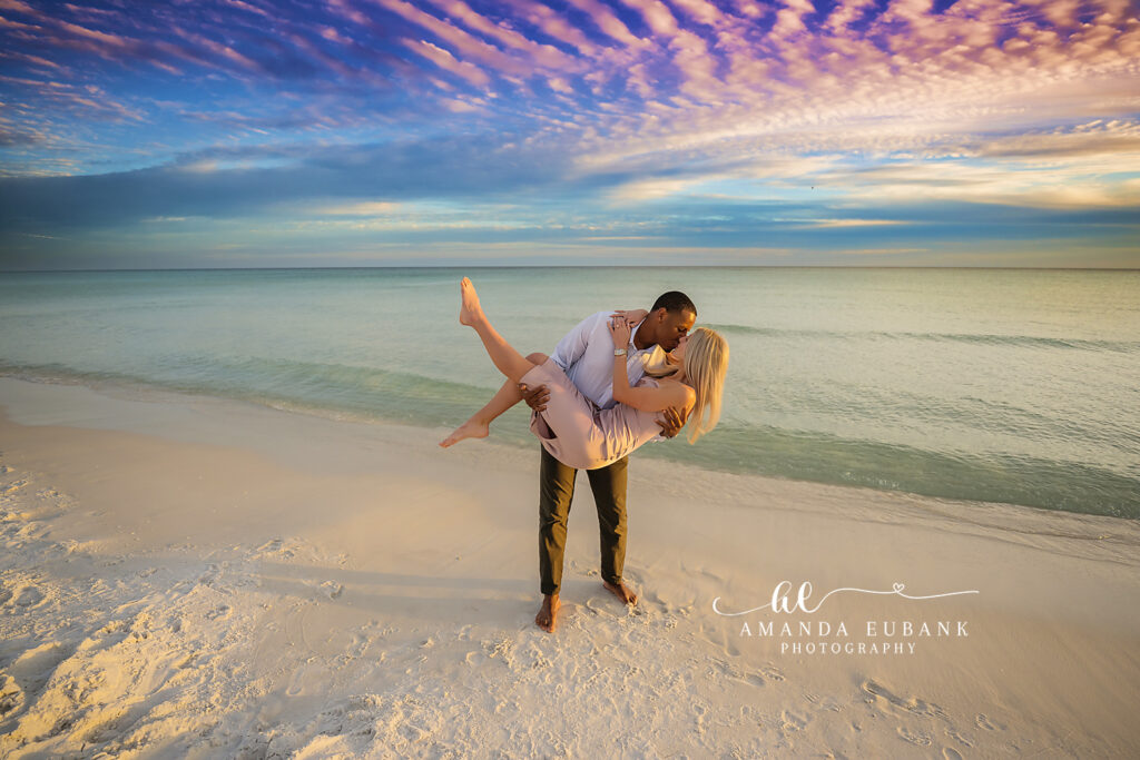 30A surprise beach proposal engagement photography: groom sweeps fiancee into a romantic barefoot dip and kiss.