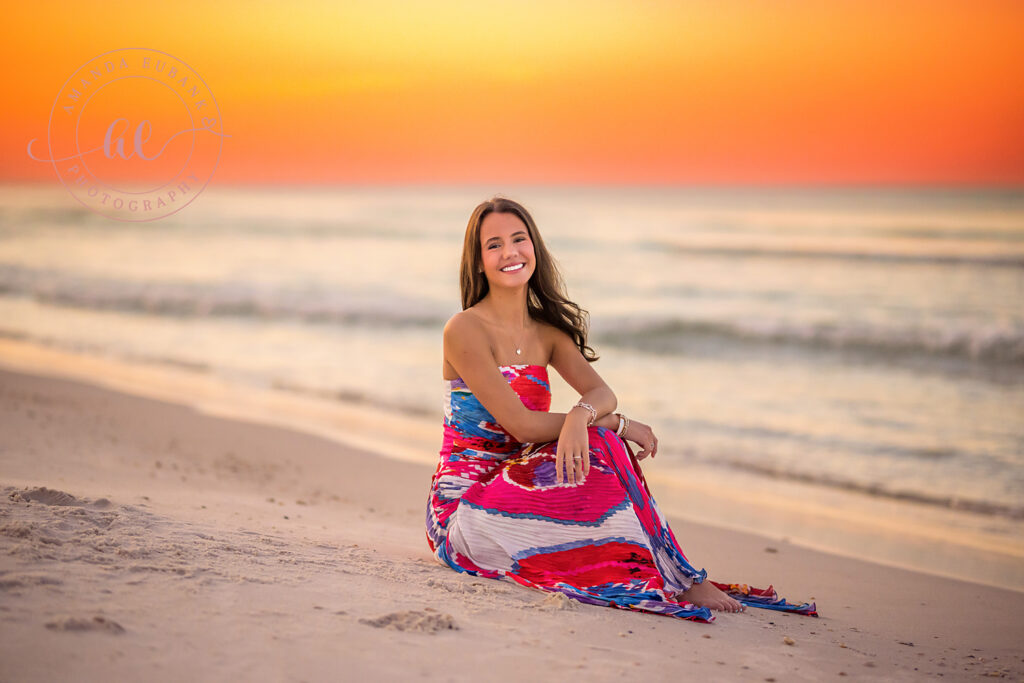 Senior Libba seated in the Seacrest sand in a tropical print maxi gown beneath a fiery orange sunrise.
