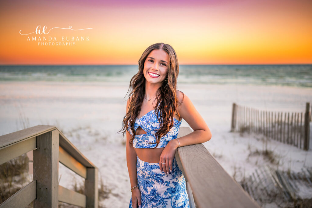 Senior girl leans on weathered boardwalk railing at 30A with dune fencing and sea oats around her, framed by a tangerine sunset glow.