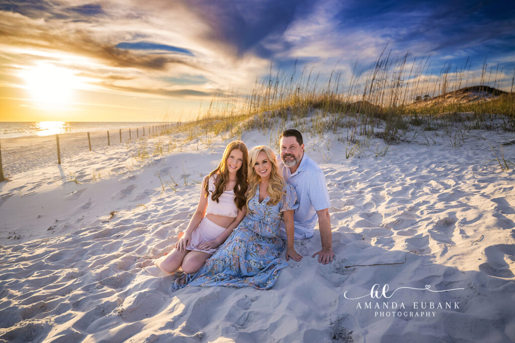 Family of three sits in the sugar-white Watercolor 30A dune sand at sunset under a painted peach and gold sky.