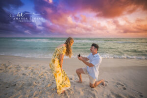Carillon Beach surprise proposal as boyfriend grins on one knee holding the open ring box up to his girlfriend.