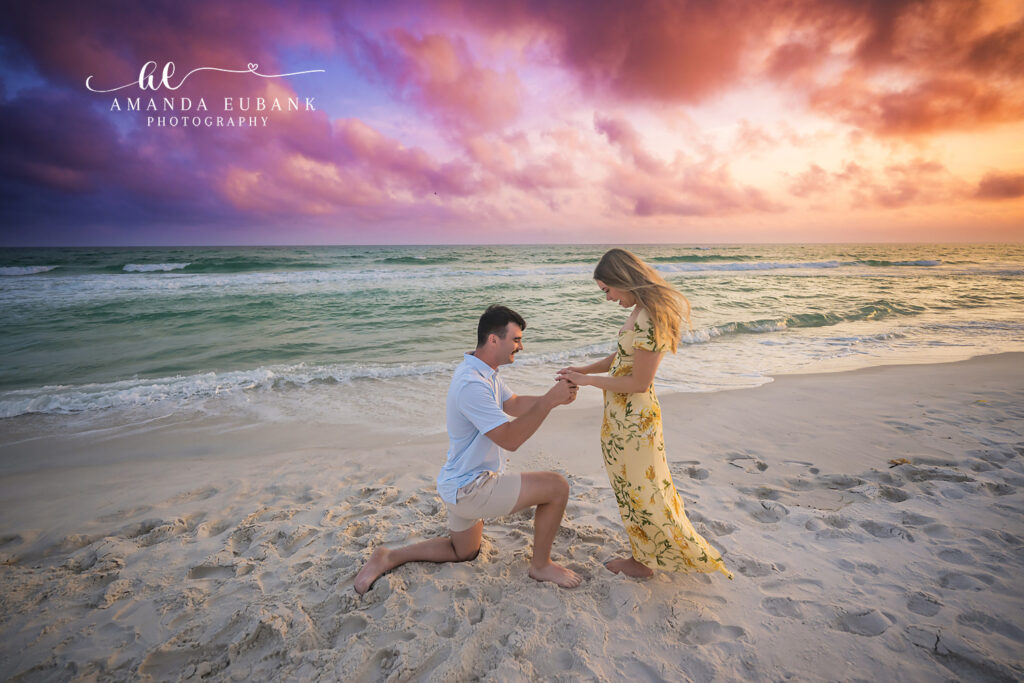 Carillon Beach engagement photography: boyfriend on one knee slides the engagement ring onto her finger at sunset.