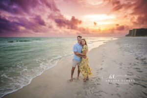 Classic Carillon Beach engagement portrait, fiance in pale blue polo and fiancee in yellow floral under a gold sky.