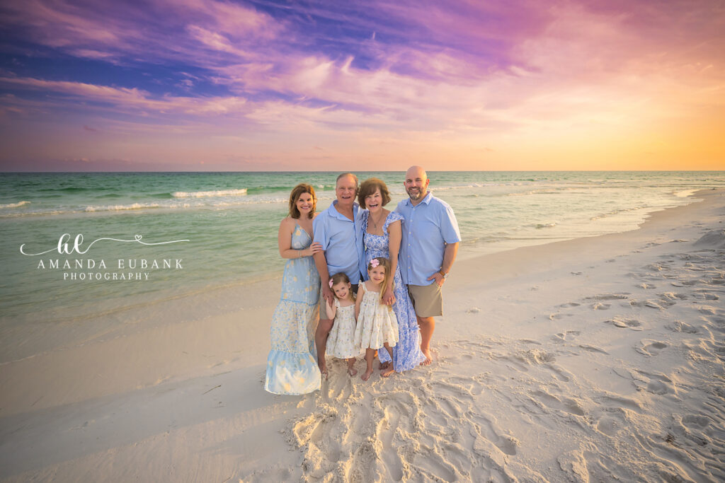 Three-generation DiLoreto family in coordinated soft blues and ivory lace stand on Destin sand at sunset with two little granddaughters in front and the emerald Gulf behind.