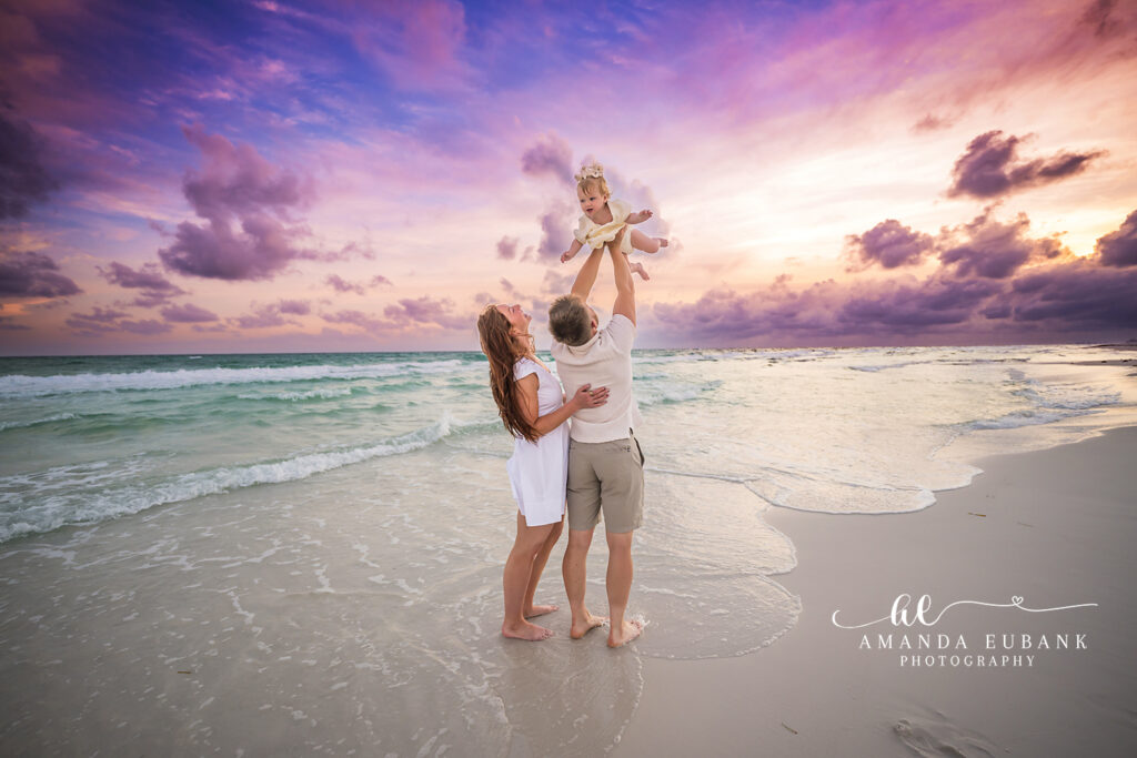 Dad lifts toddler in butter-yellow dress overhead while mom in white watches and smiles along Watercolor 30A Beach beneath a lavender sunset.