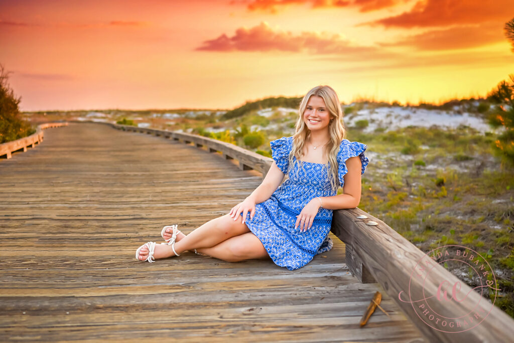 Senior Sophia seated on the wooden Watersound Beach Club dune boardwalk leaning against the railing in a tiered blue sundress at fiery orange and peach Florida sunset