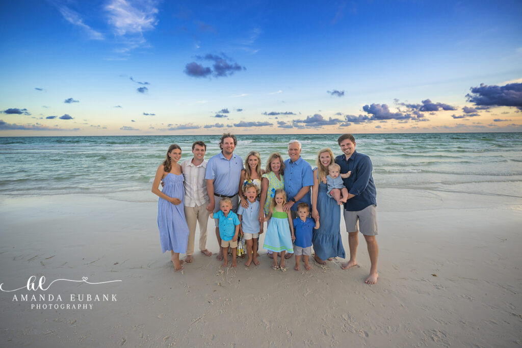 Large extended Martin family of fourteen stands together on the Watercolor, FL shoreline at sunset, all coordinated in shades of blue and green.