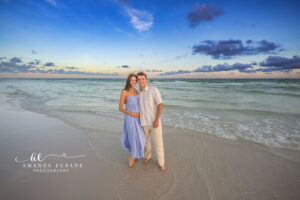 Young couple stand together at the Watercolor, FL sunset shoreline, woman in a flowy cornflower blue maxi and man in cream linen with the Gulf surf rolling behind them.