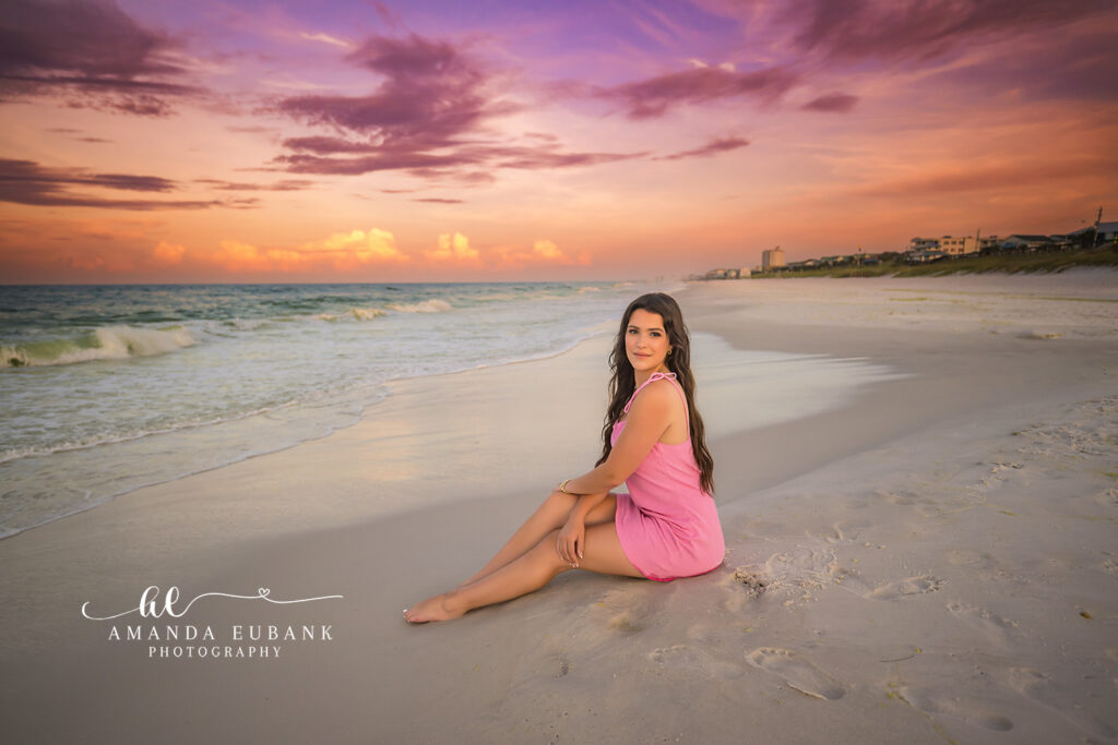 Senior Anna seated on the sand with legs crossed in a pink mini dress against a dramatic purple pink and orange streaked sunrise sky over the Destin shoreline