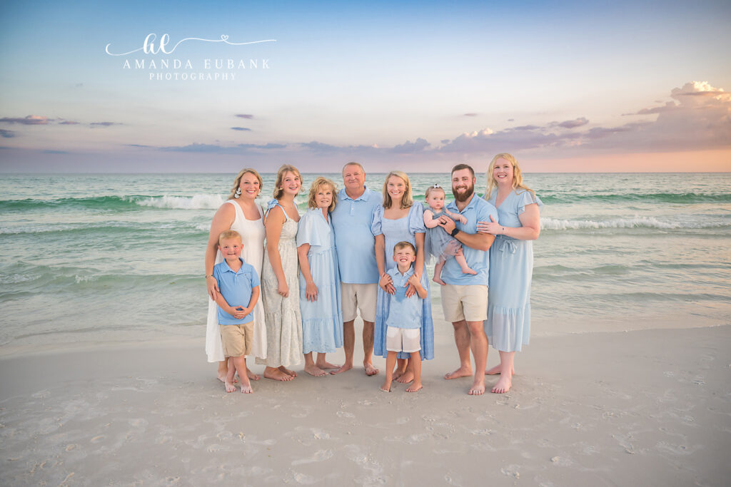 Extended family of eleven coordinated in light blue and white linen stands together on the Destin, FL shoreline at sunset, gentle Gulf surf rolling behind.