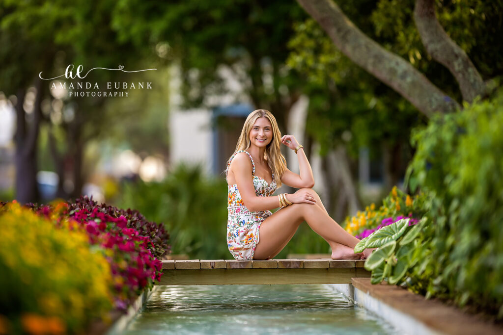 High school senior in a colorful floral mini dress sits barefoot on the wooden ledge of the Watercolor, FL lily pond surrounded by magenta and yellow blooms.