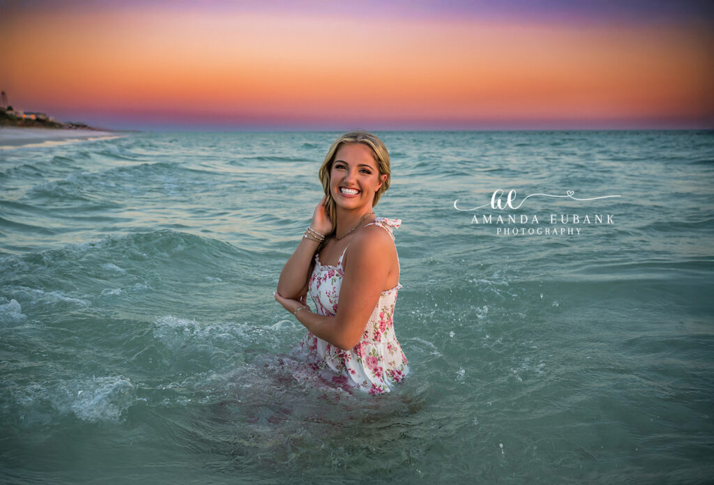 Watercolor 30A senior session: senior in a pink floral sundress wading waist-deep in the Gulf at post-sunset glow.