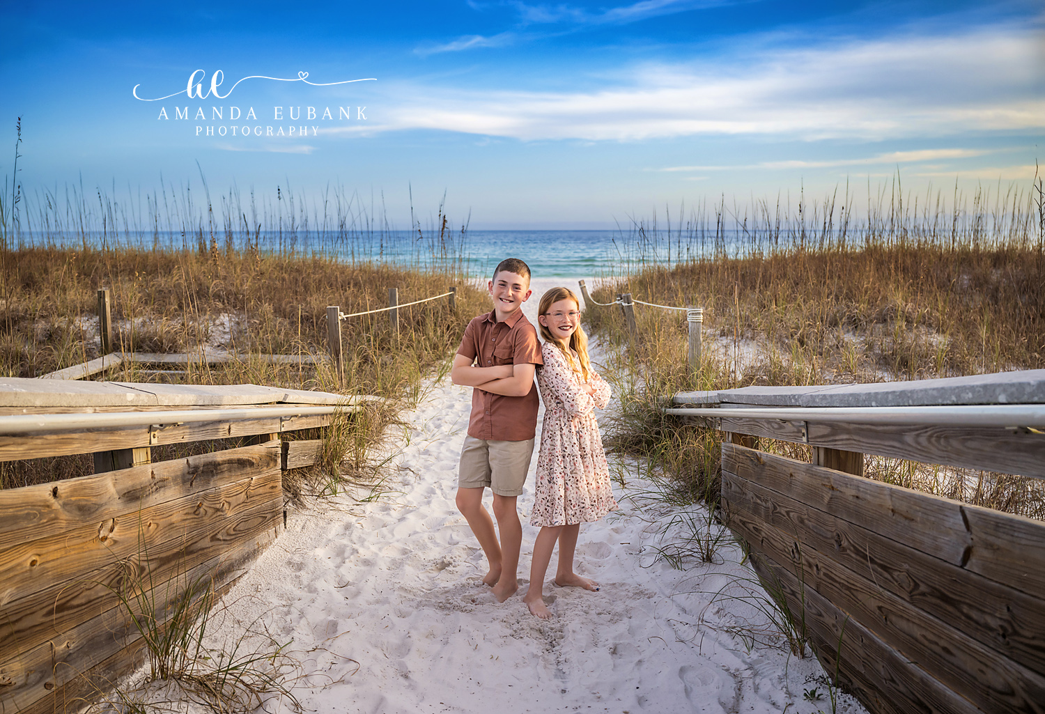 Henderson Beach State Park Boardwalk Sibling Portrait | Destin FL Family Photographer Brother in a rust polo and sister in a floral dress standing arm in arm on the wooden boardwalk between sea oat dunes at Henderson Beach State Park in Destin FL