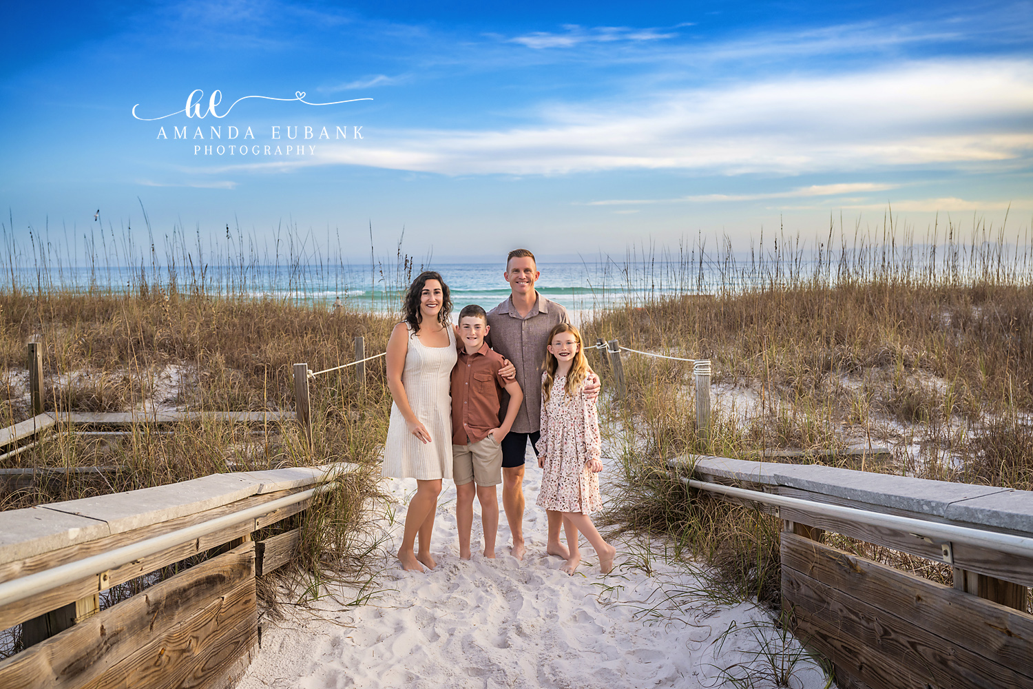 Henderson Beach State Park Family of Four Portrait | Destin FL Family Photographer Family of four standing together on the wooden boardwalk between sea oat dunes at Henderson Beach State Park in Destin FL with the Gulf and a blue sky behind them