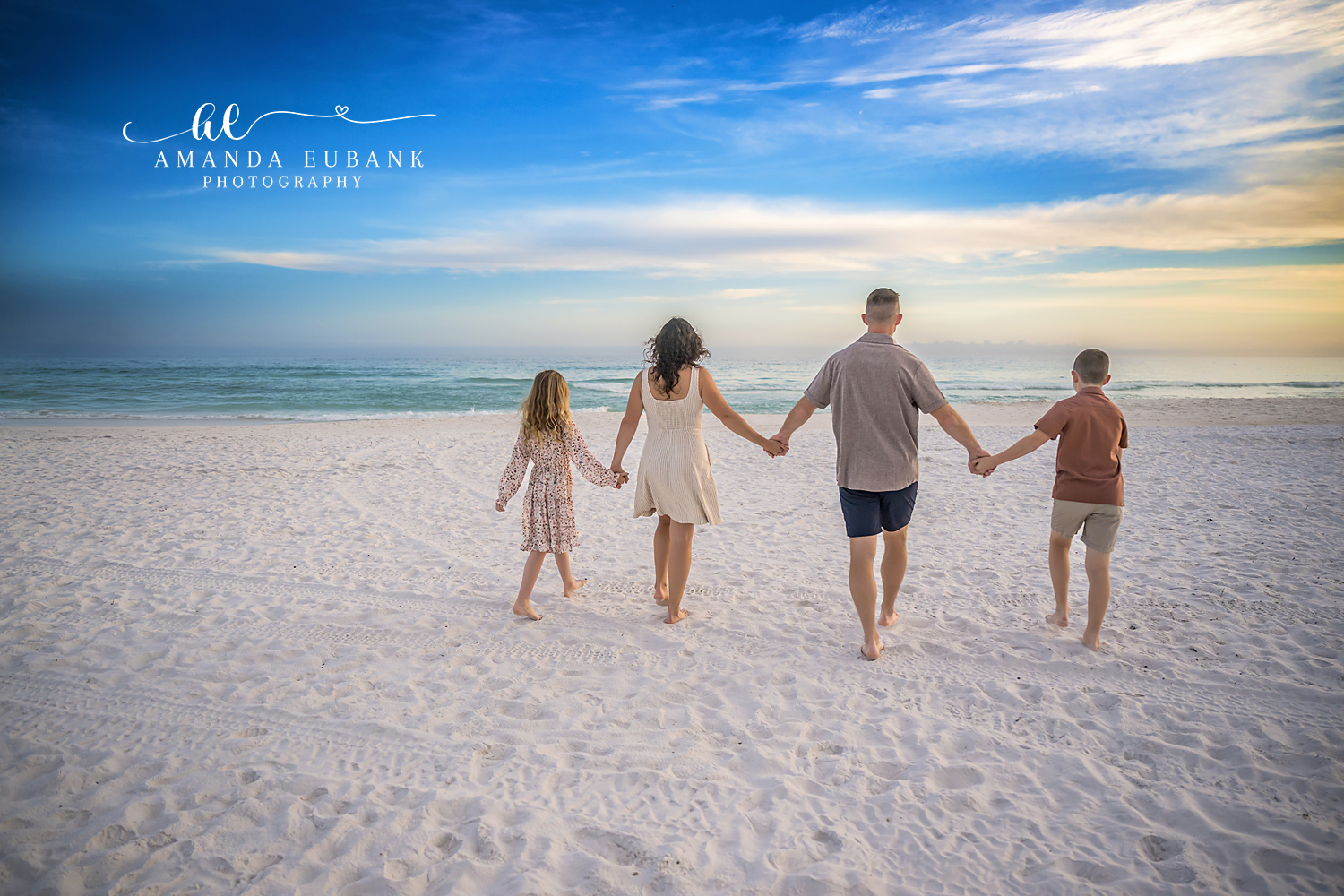 Destin Family Walking Hand in Hand to the Gulf | Destin FL Family Photographer Family of four walking hand in hand away from camera toward the Gulf at Henderson Beach State Park in Destin FL beneath a soft blue sunset sky