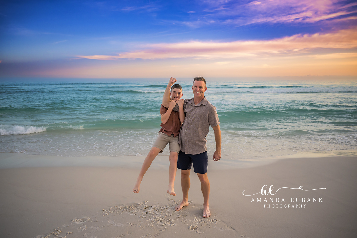 Destin Father and Son Sunset Beach Portrait | Destin FL Family Photographer Father in a sage shirt and son in a rust polo laughing together with son flexing on the Destin FL beach at sunset with the Gulf and a pastel sky behind