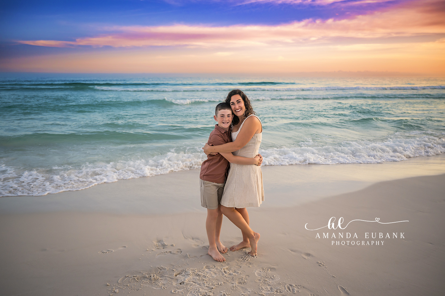 Destin Mother and Son Sunset Hug Portrait | Destin FL Family Photographer Mom in a cream sundress hugging her son in a rust polo on the Destin FL beach at sunset with the Gulf surf and a pastel pink and orange sky